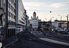 Kuvassa on kaupunkikuva, jossa näkyy leveä katu, suojatie, rakennuksia ja katuvaloja.